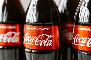 Close-up van glazen frisdrank Coca-Cola-flessen met het herkenbare rode etiket en witte "Original Taste"-tekst, scherp in beeld tegen een wazige achtergrond van andere flessen. Experts waarschuwen: frisdrank maakt ons zieker dan we denken